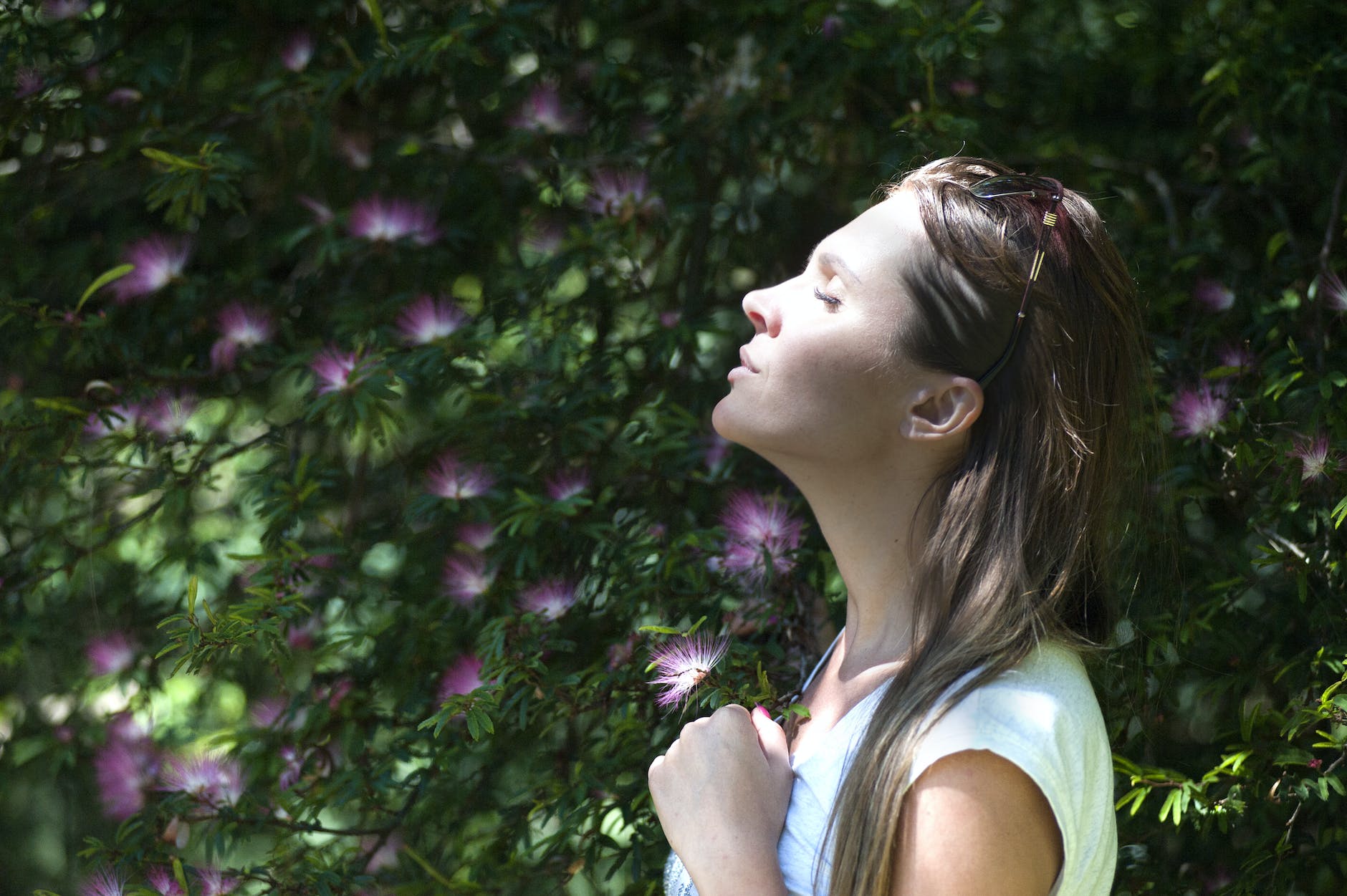 Woman closing her eyes against sun light standing near purple petaled flower plant anger